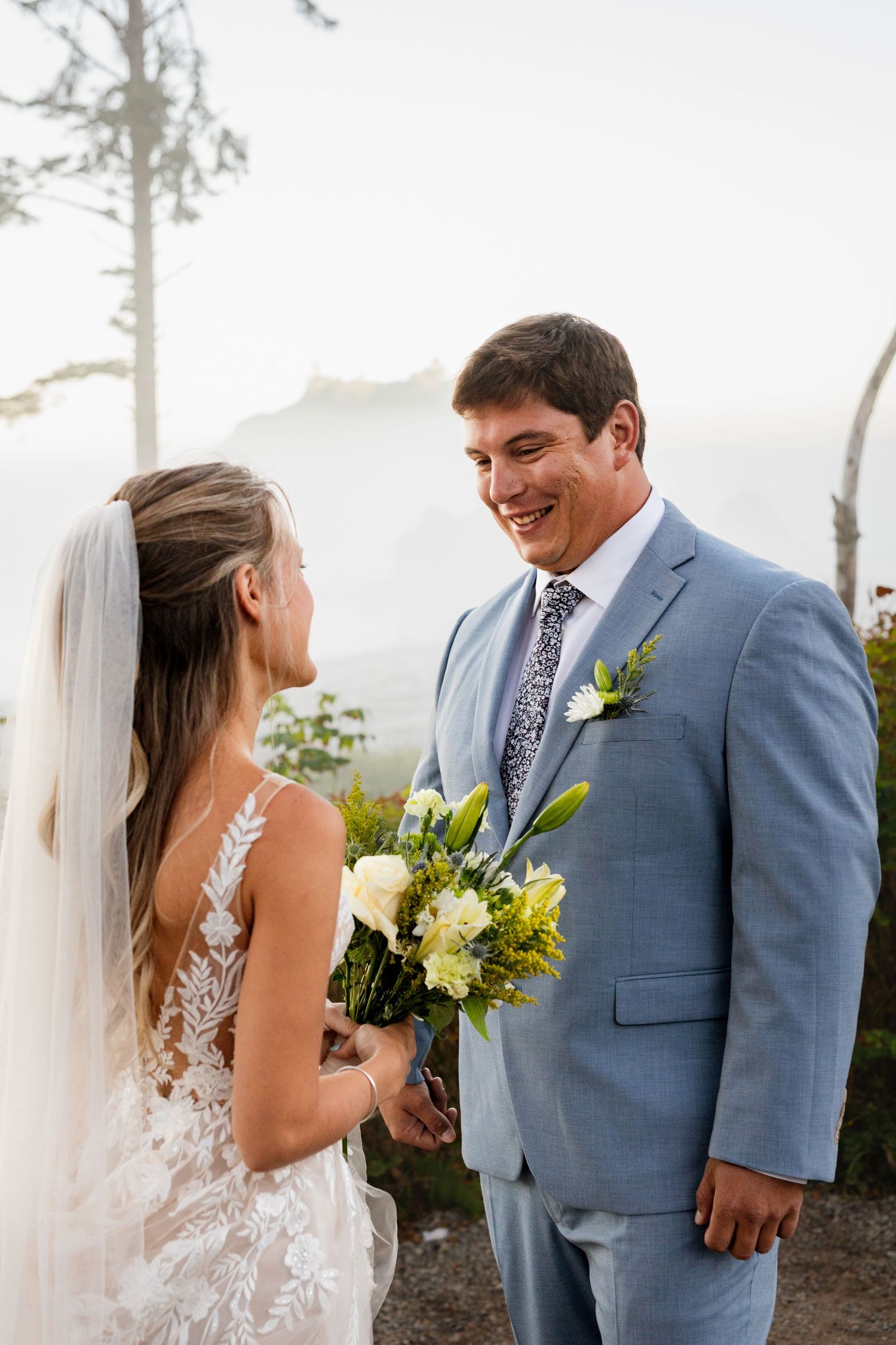 Beaming groom in light blue suit holds bouquet during first look with lace-gowned bride on coastal bluff.