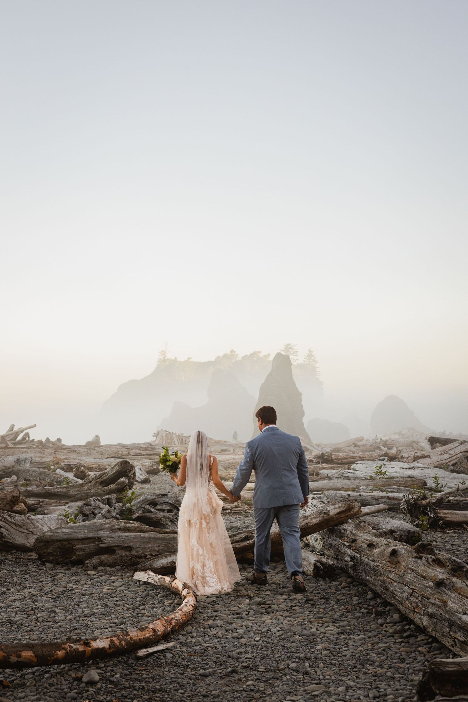 Wedding couple holding hands facing misty sea stacks on a driftwood-covered beach.