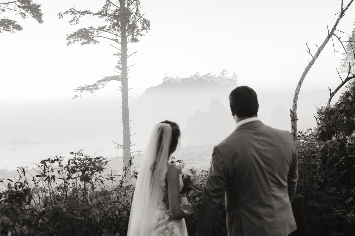 Black and white photo of bride and groom gazing at misty sea stack coastline from forested overlook.