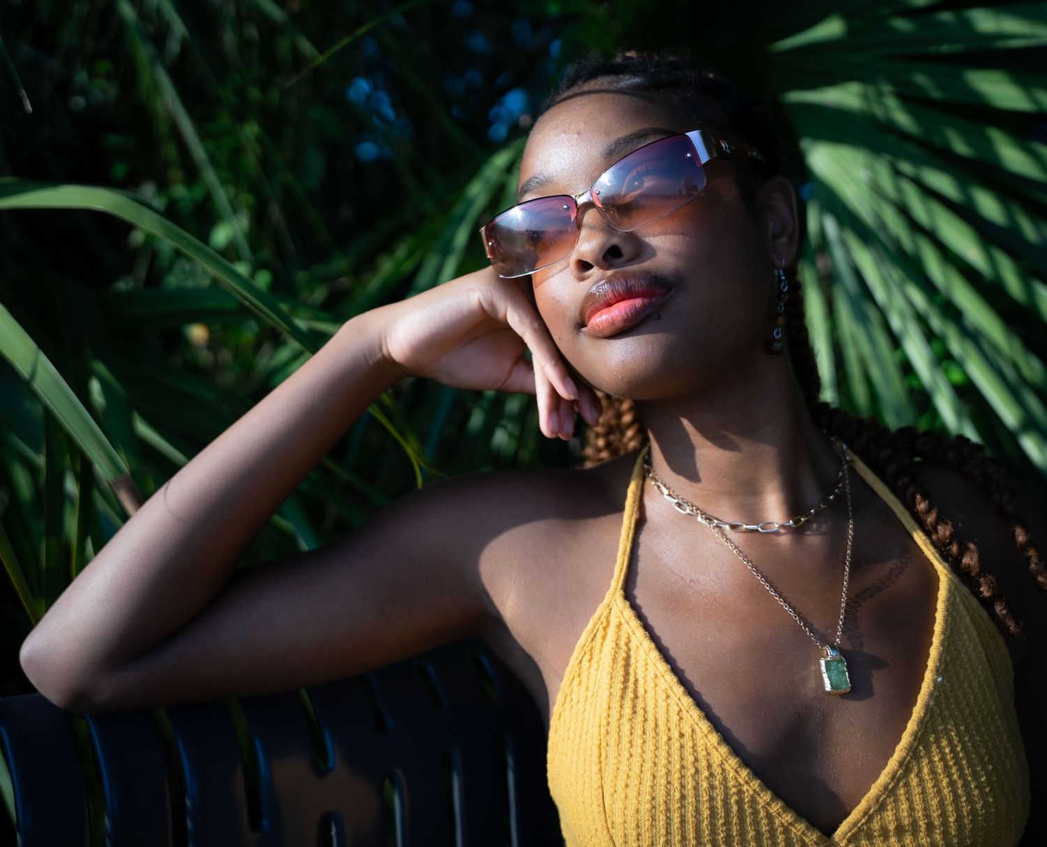Person in a yellow knit top poses stylishly against a backdrop of green palm leaves in natural lighting.