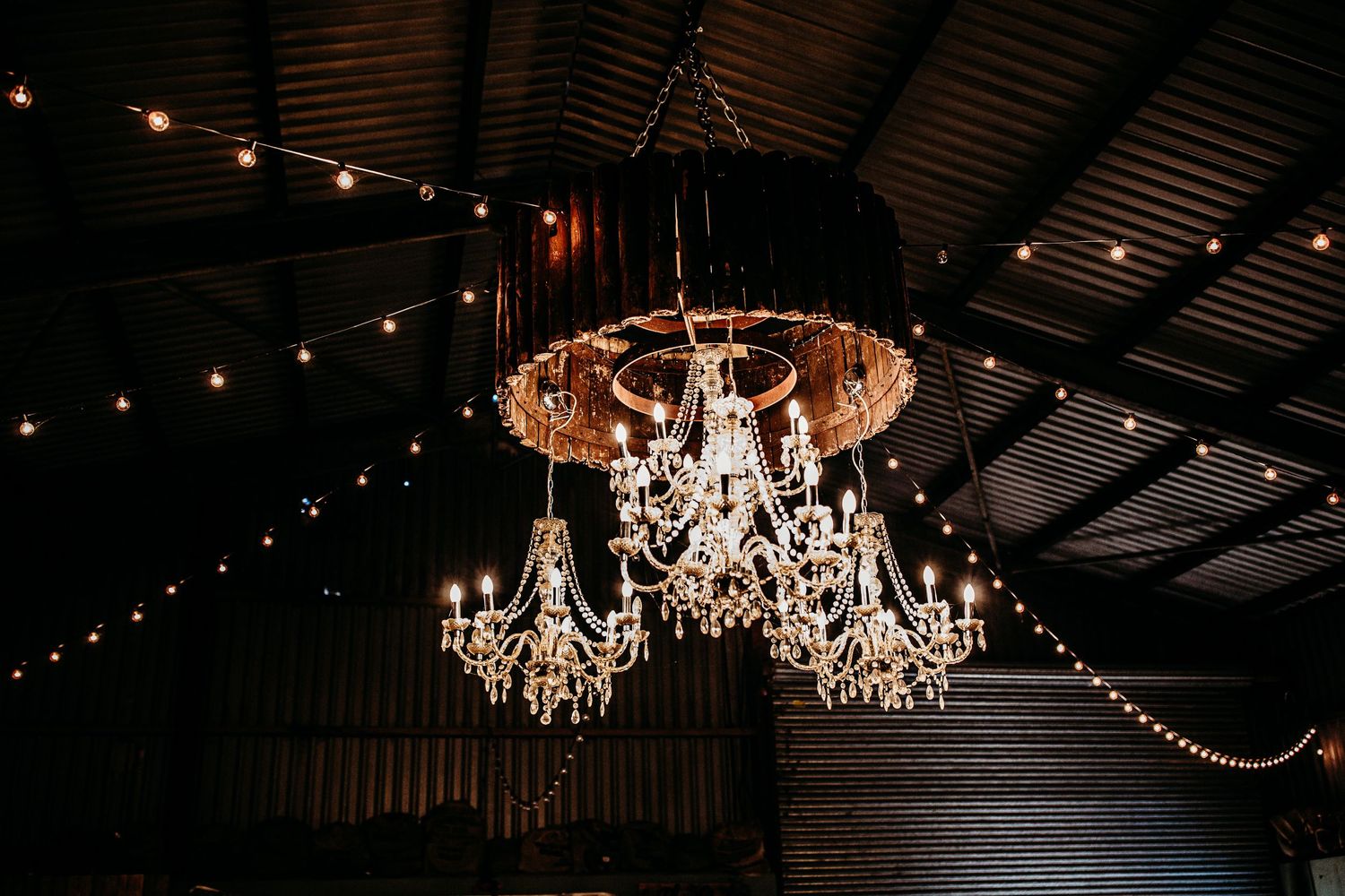 Elegant crystal chandeliers and string lights hanging from rustic barn ceiling.