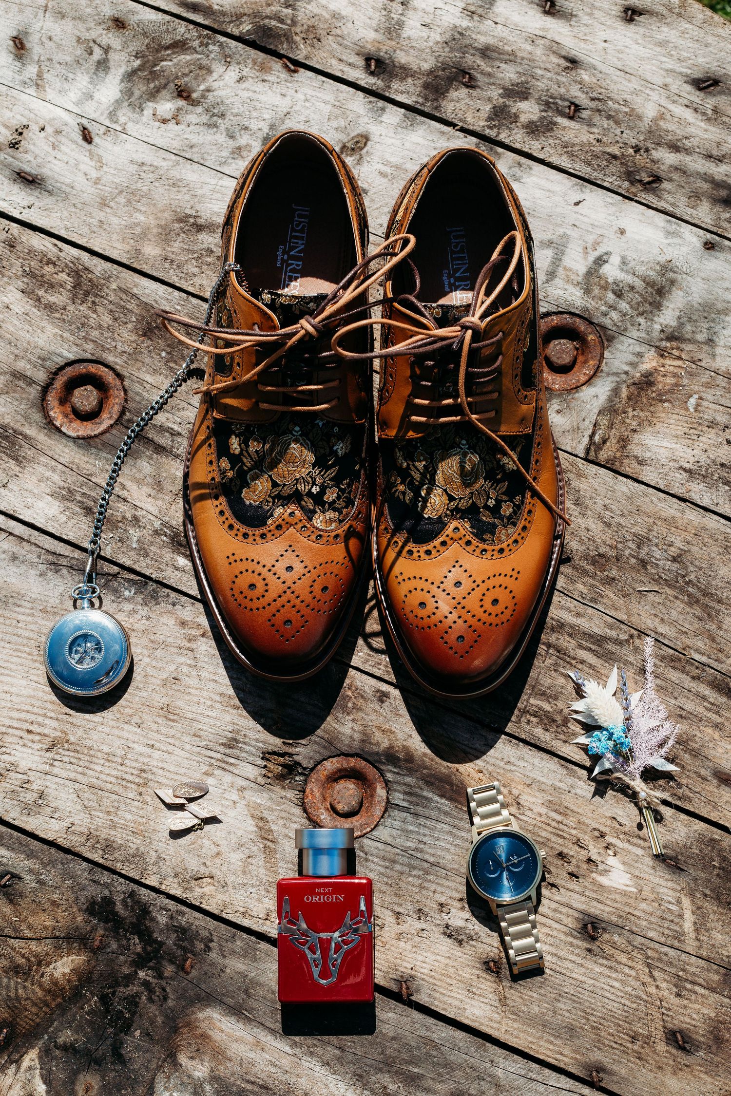 Brown leather wingtip oxford shoes with accessories on rustic wooden surface.