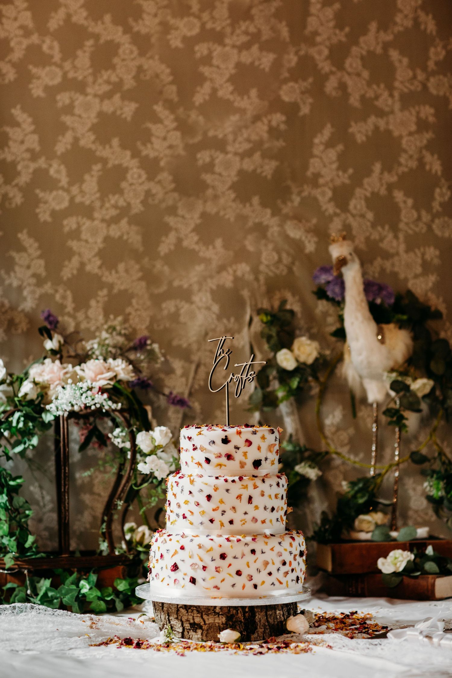Three-tiered white wedding cake with colorful floral decorations on rustic wooden stand.