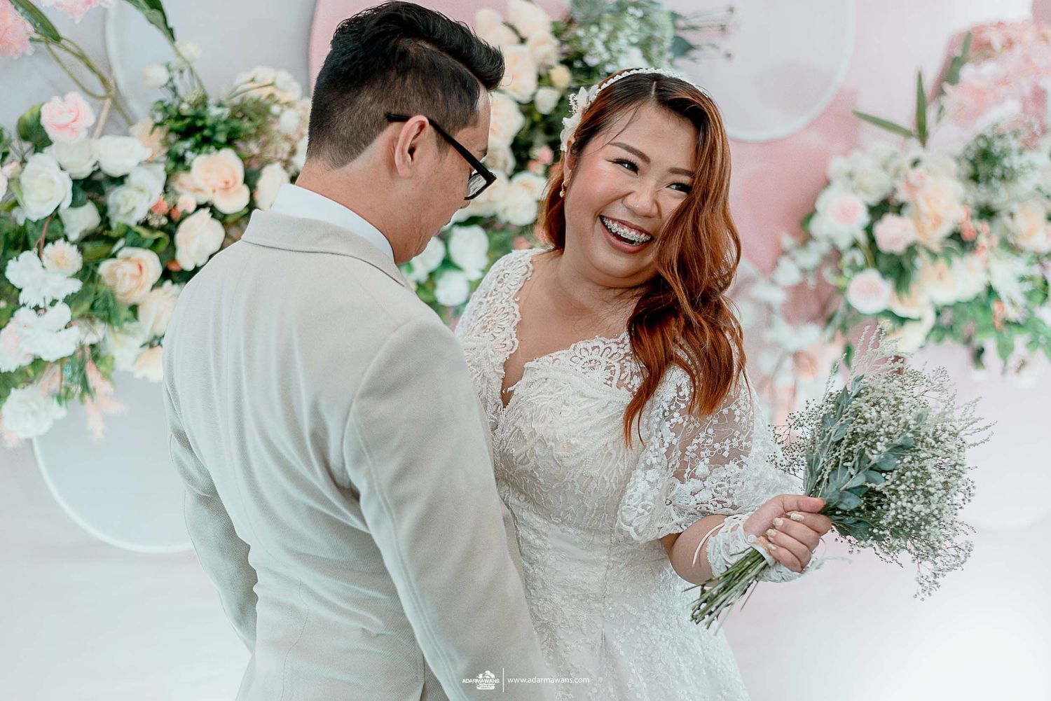 Joyful wedding couple laughing together surrounded by pink and white floral decorations.