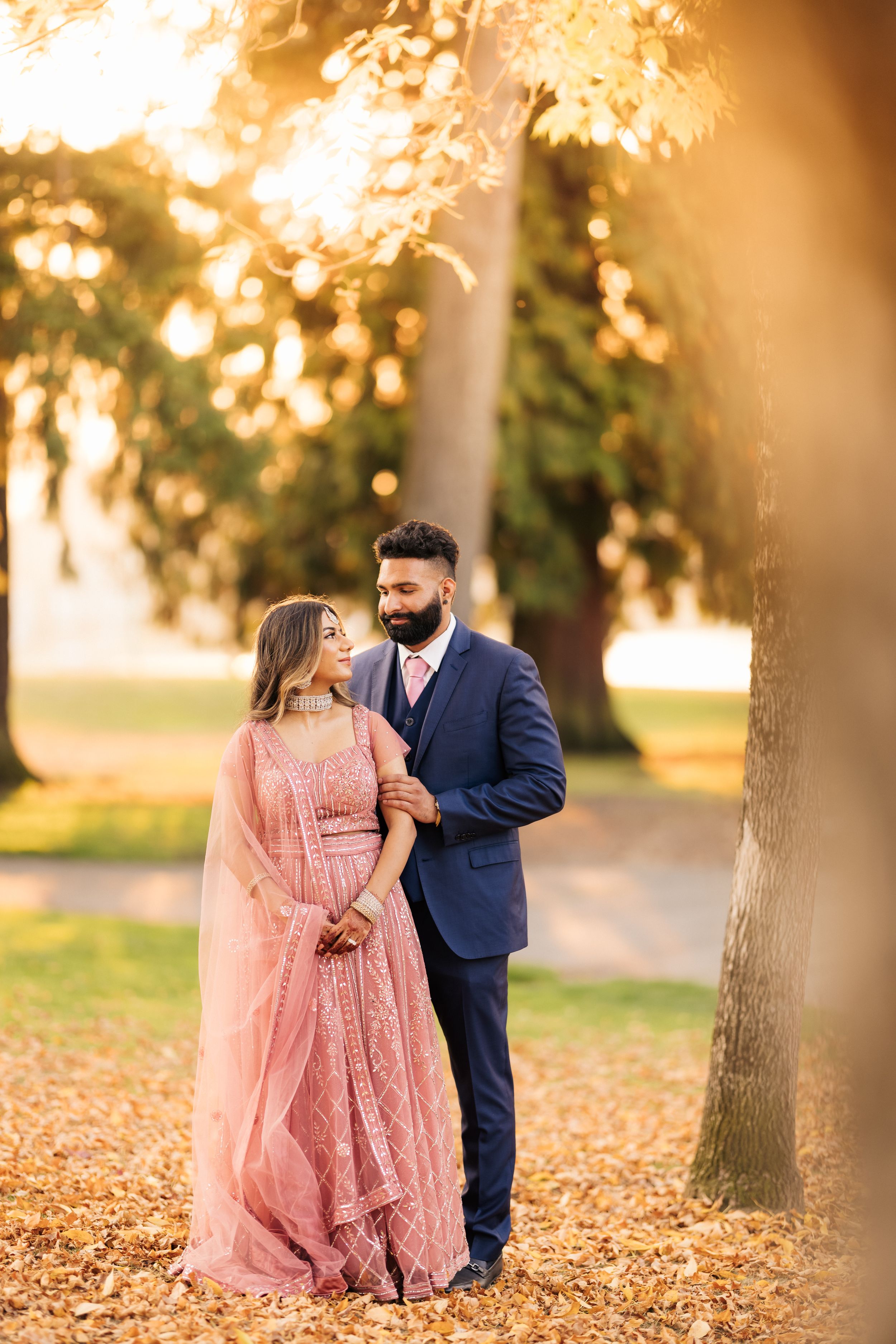 Couple in formal attire embracing in a park during golden hour with autumn leaves on the ground.