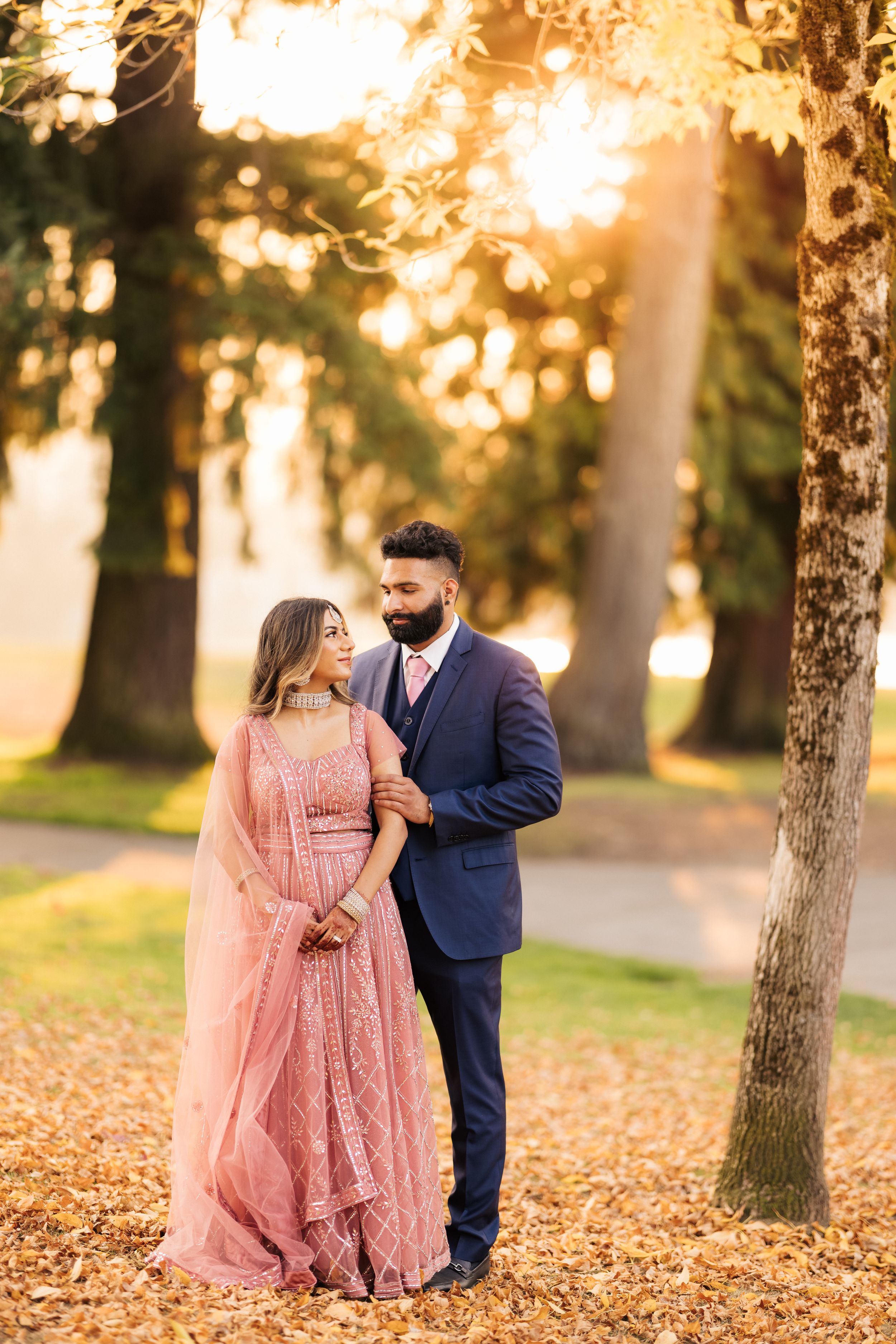 A couple poses beneath fall trees with warm sunset light filtering through branches.