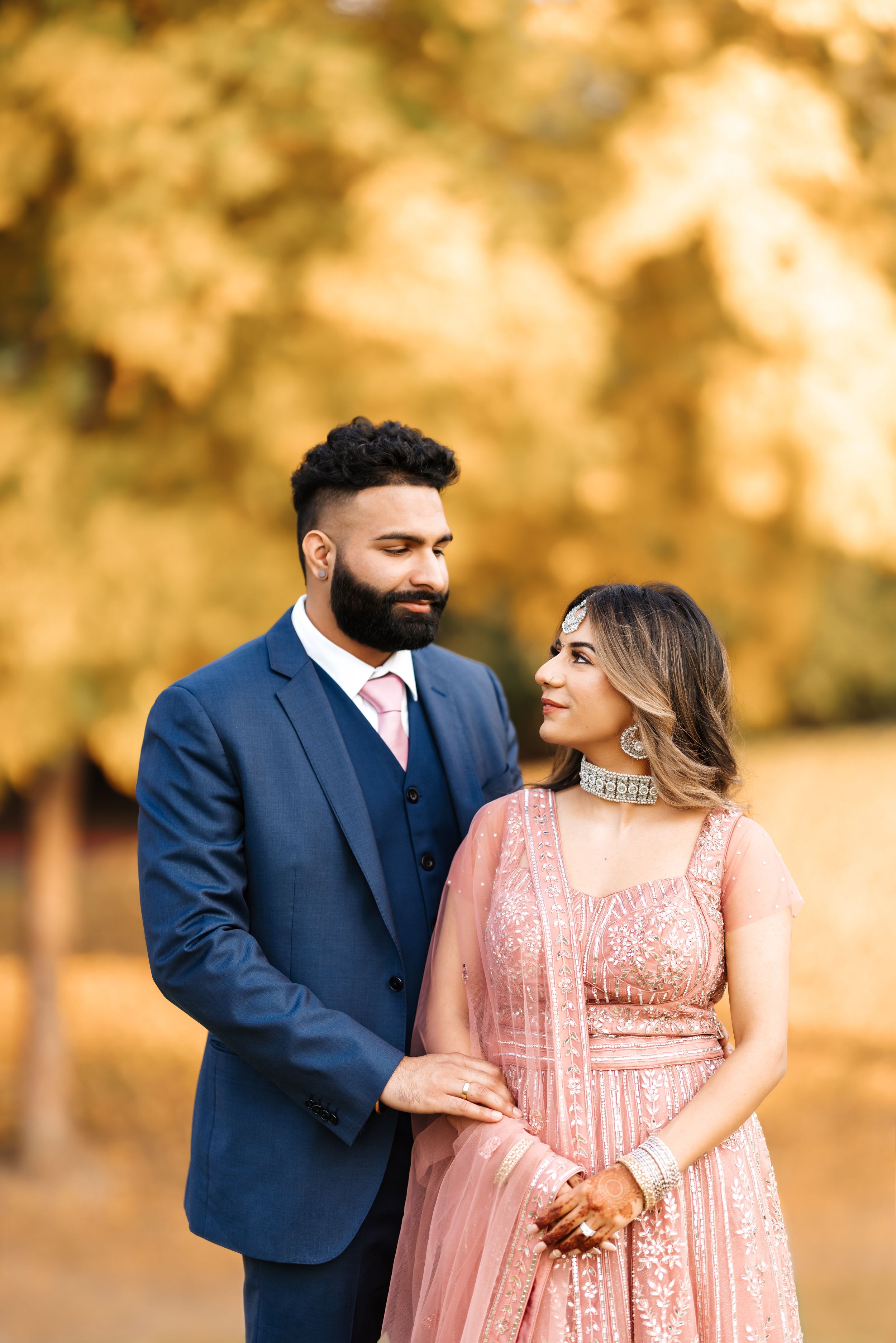 Elegant couple in navy suit and pink gown pose together against autumn foliage.