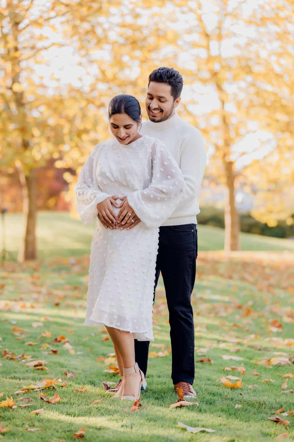A couple in white outfits poses together on grass with fall trees in the background.
