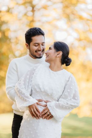 A couple in matching white sweaters shares a tender moment against golden autumn foliage.