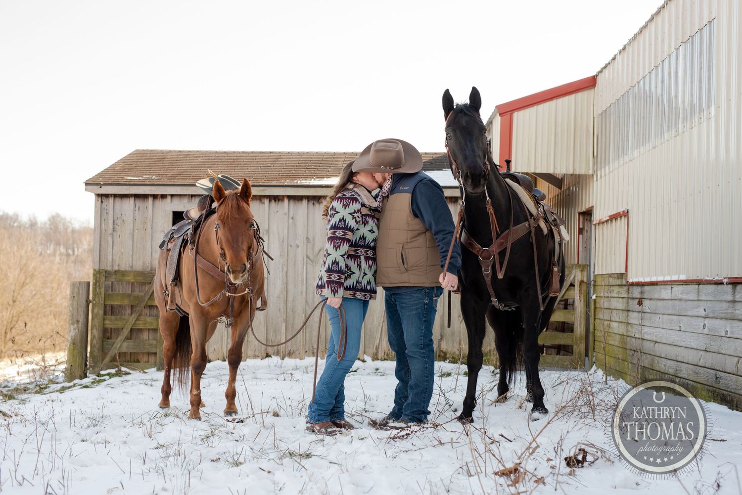 Two horses stand with riders in front of rural buildings on a snowy winter day.