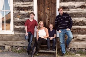 Family of four poses on wooden bench against log cabin wall.