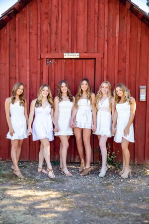 Group in white dresses lined up against red barn exterior.