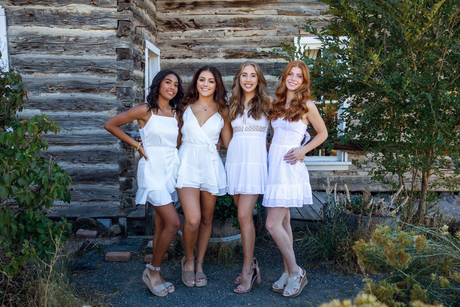 Four friends in white dresses pose against rustic log cabin wall.