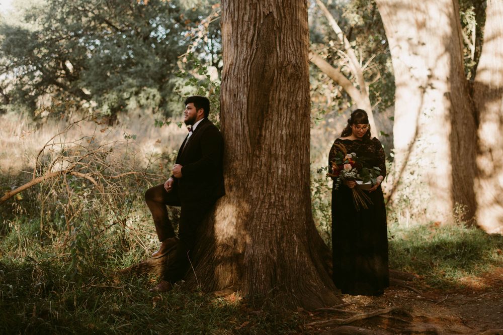Two people in formal attire posing beside large trees in a romantic forest setting.