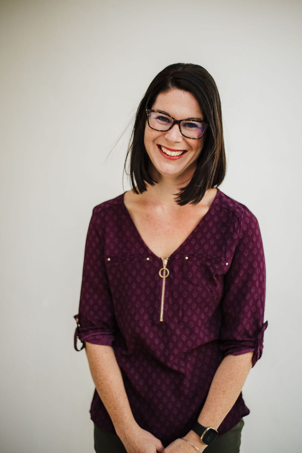 Professional wearing glasses and a burgundy patterned top smiles warmly against neutral background.