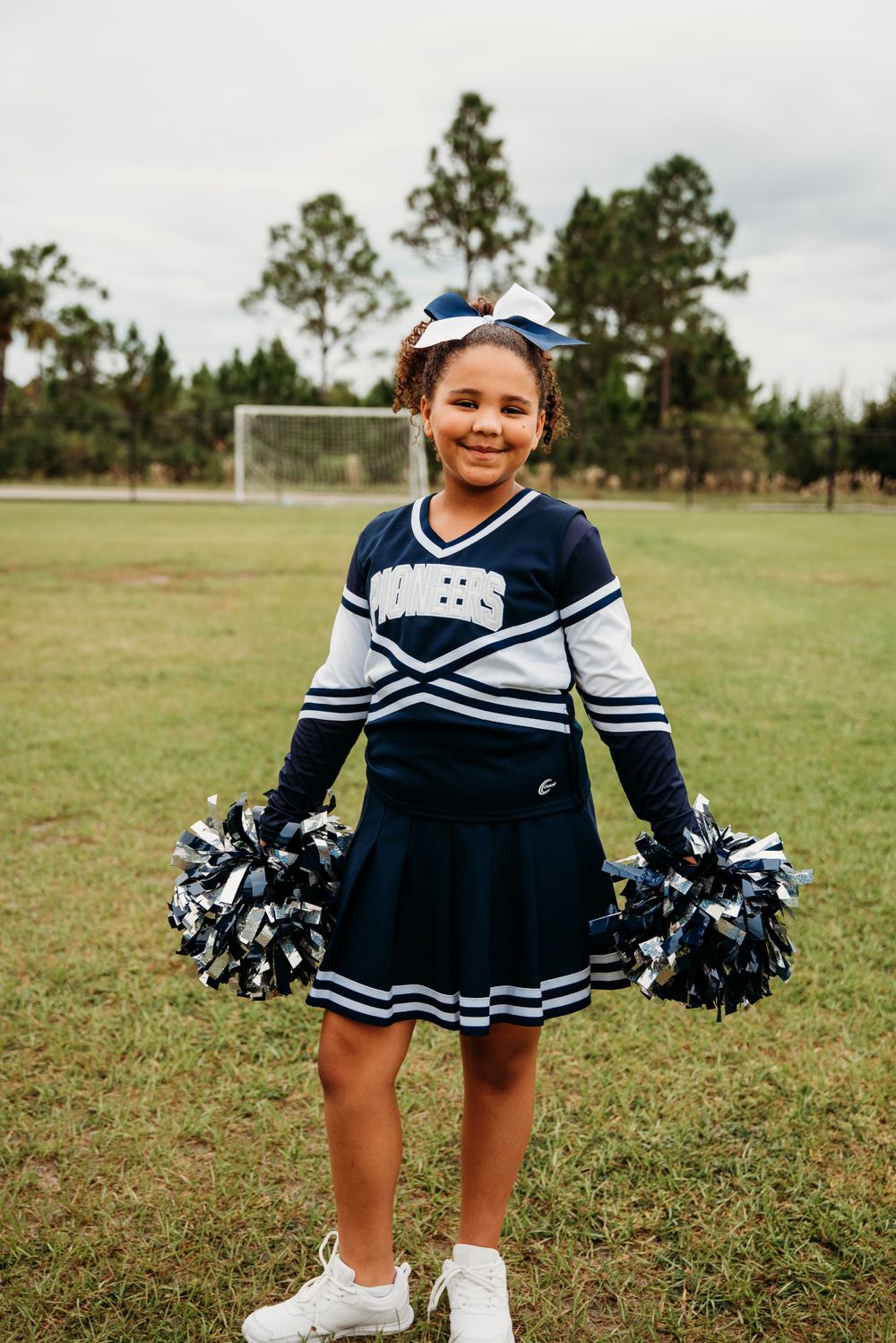 Cheerleader in navy and white uniform holding pom poms poses on sports field.