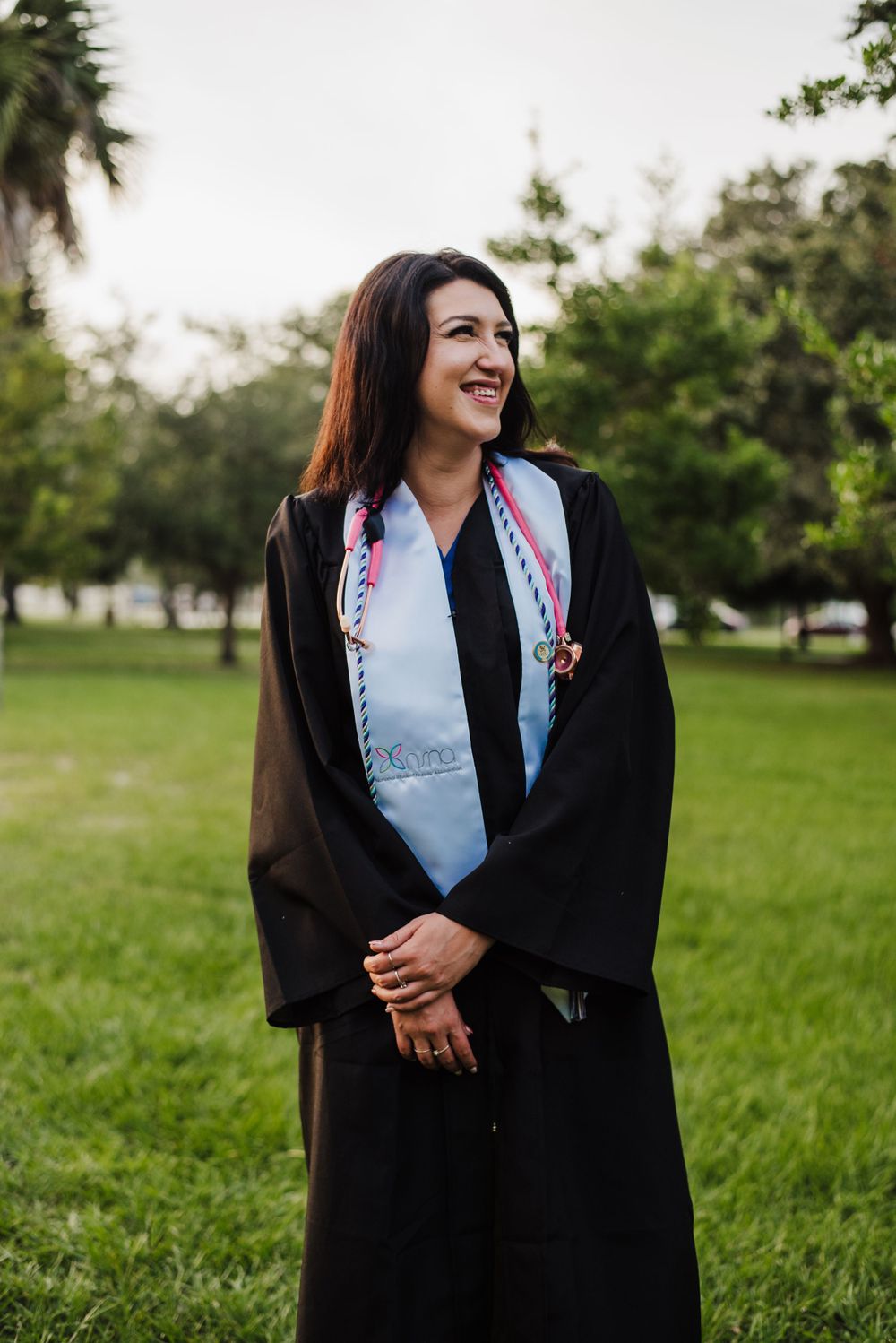 Graduate in traditional black cap and gown smiles outdoors on grassy campus.
