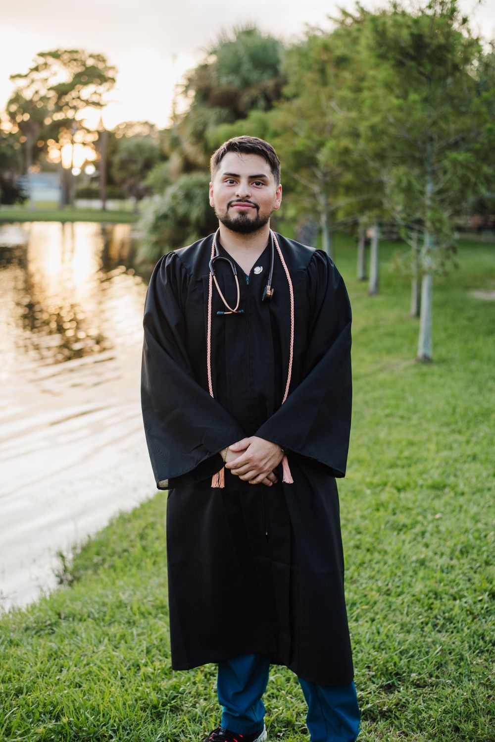 Graduate in traditional black regalia poses by waterfront at sunset.