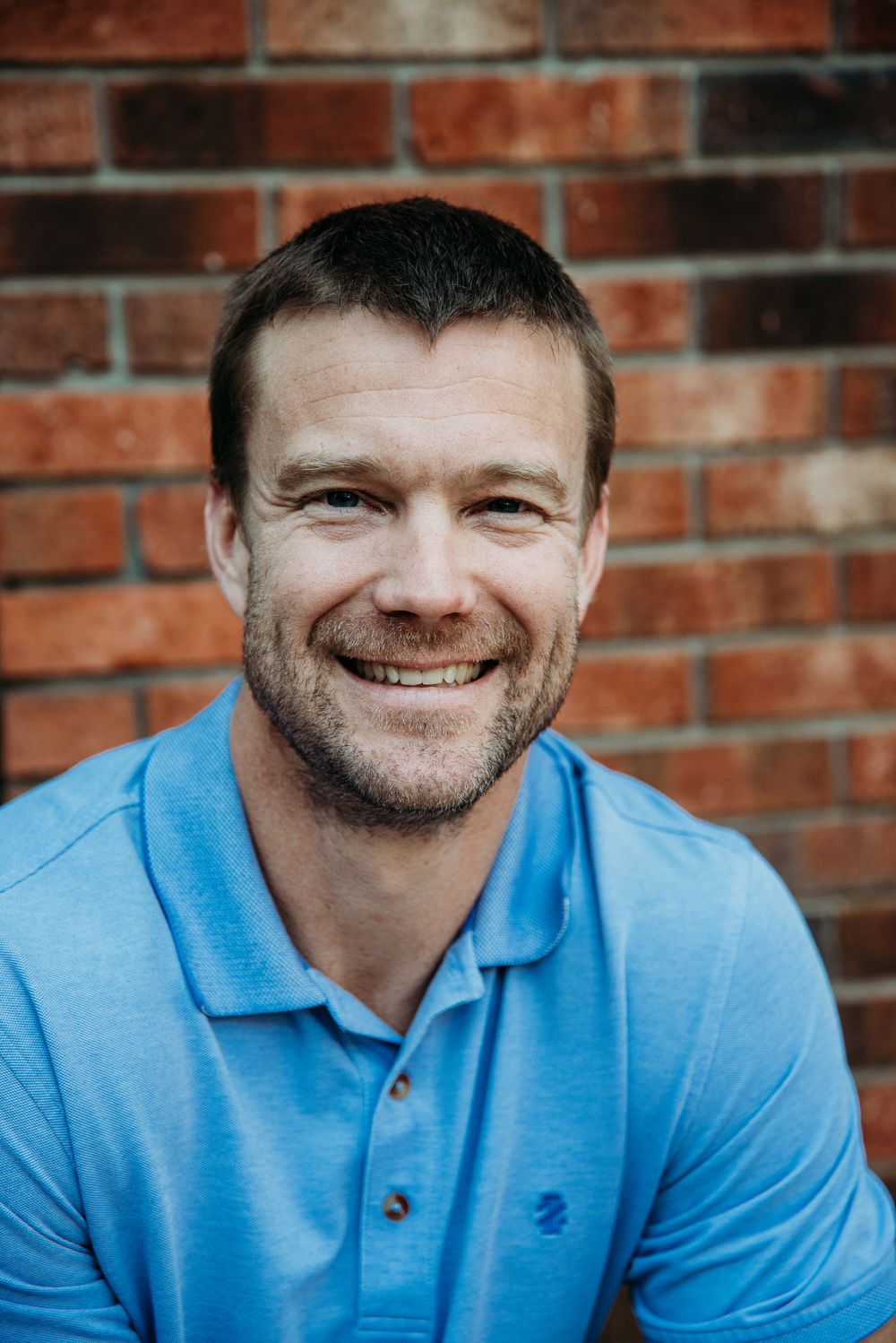 Professional headshot of person in light blue polo shirt against brick wall background.