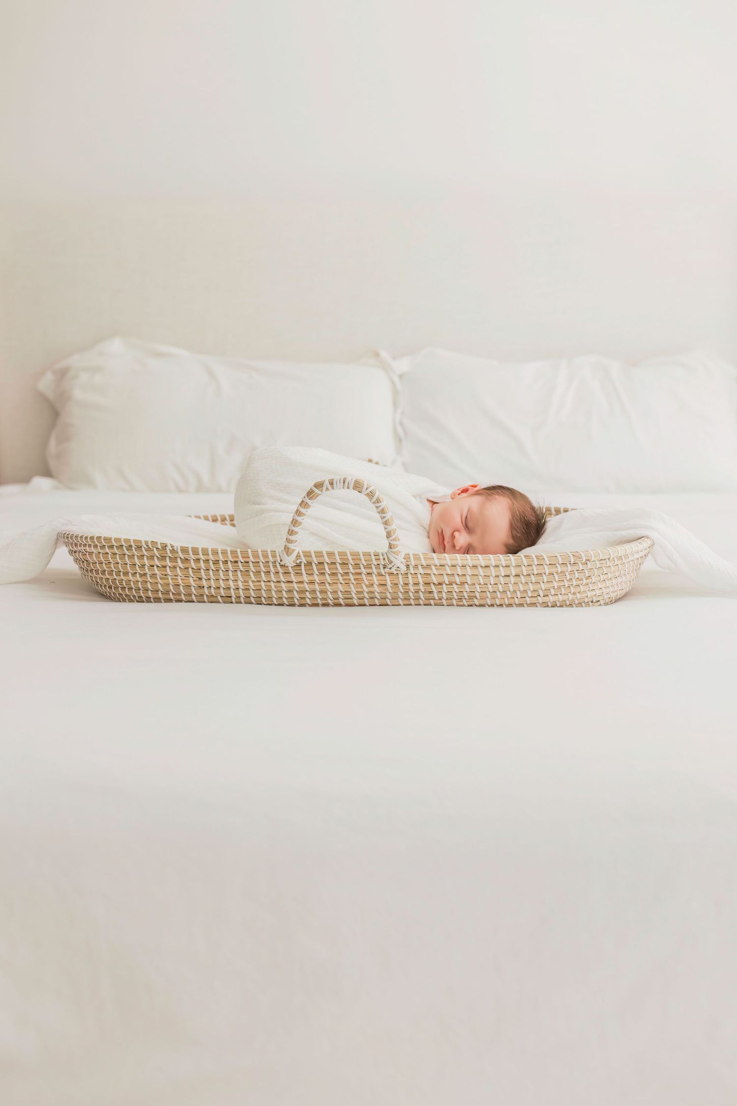Sleeping newborn swaddled in white resting in woven bassinet on bed.