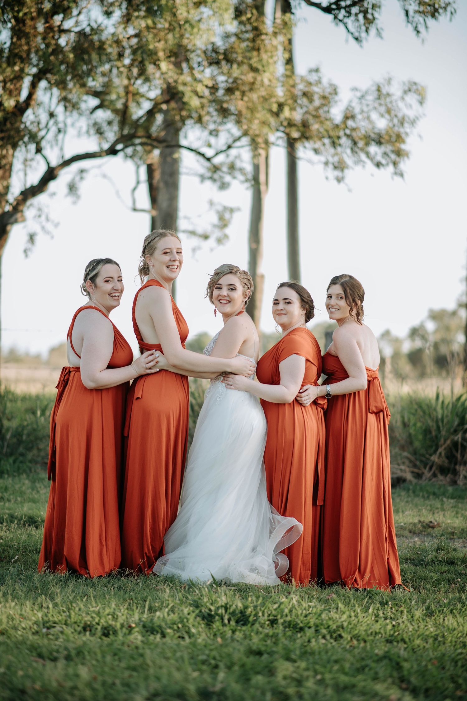 Bridesmaids in flowing orange dresses pose with a bride in white against a natural backdrop.