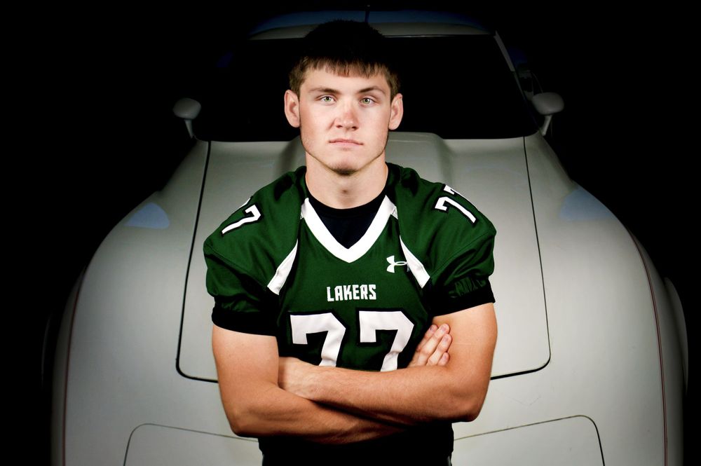 High school athlete in green football jersey number 77 poses against car hood.