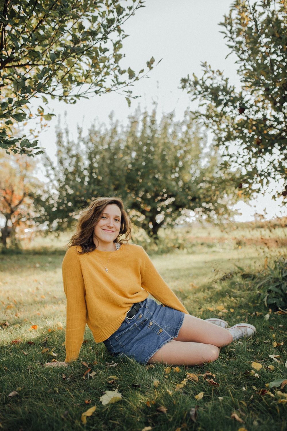 Someone in a mustard sweater and denim skirt relaxes on grass surrounded by autumn foliage.