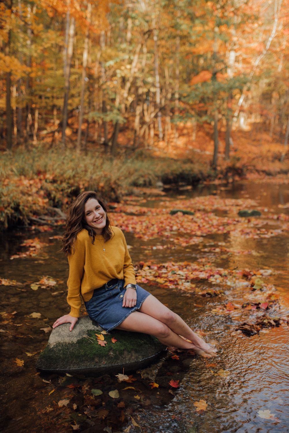 Someone in mustard sweater sits on rock by stream surrounded by autumn leaves.