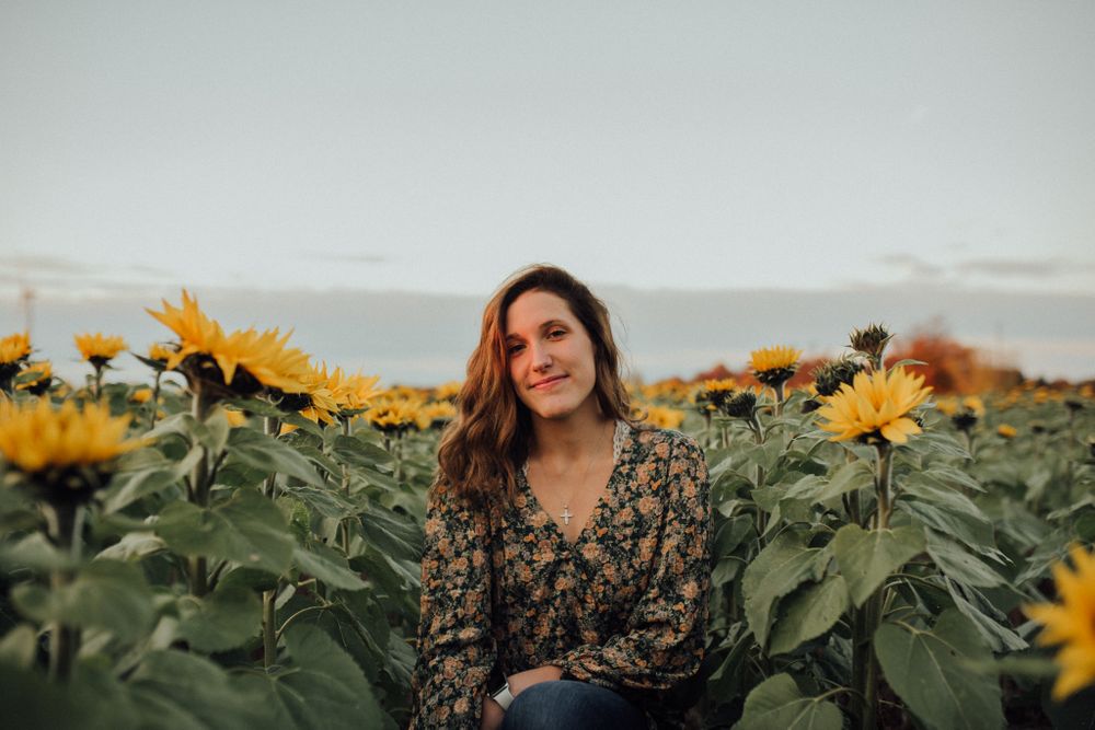 Someone sits among bright yellow sunflowers wearing a patterned dark top during golden hour.