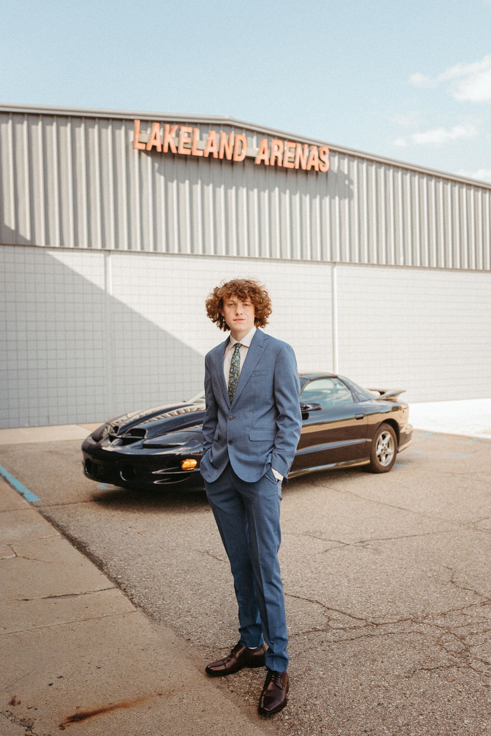 A portrait of someone in a blue suit standing in front of a black car outside a commercial building.