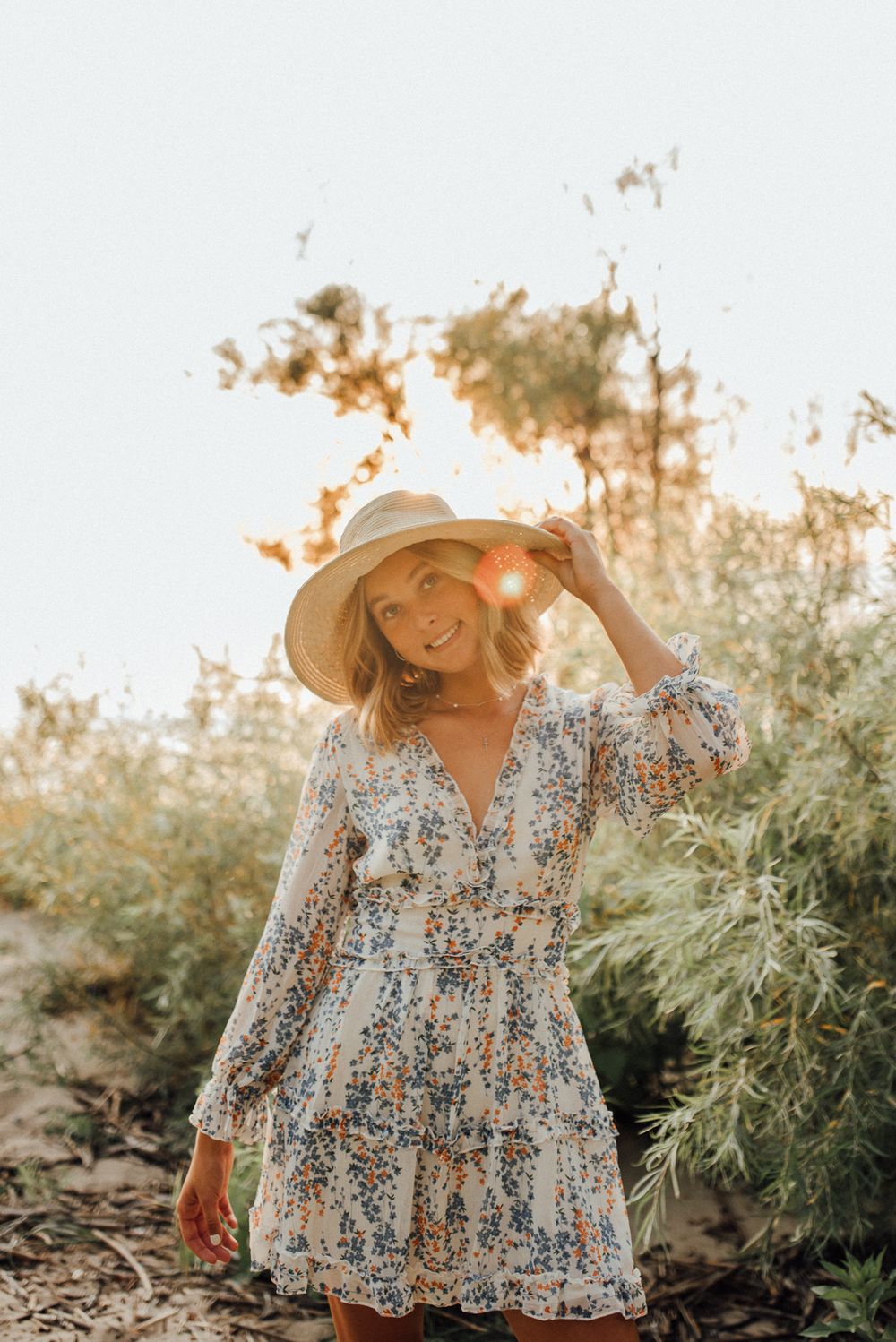 Sun-drenched portrait in a floral dress and wide-brim hat amidst natural foliage.