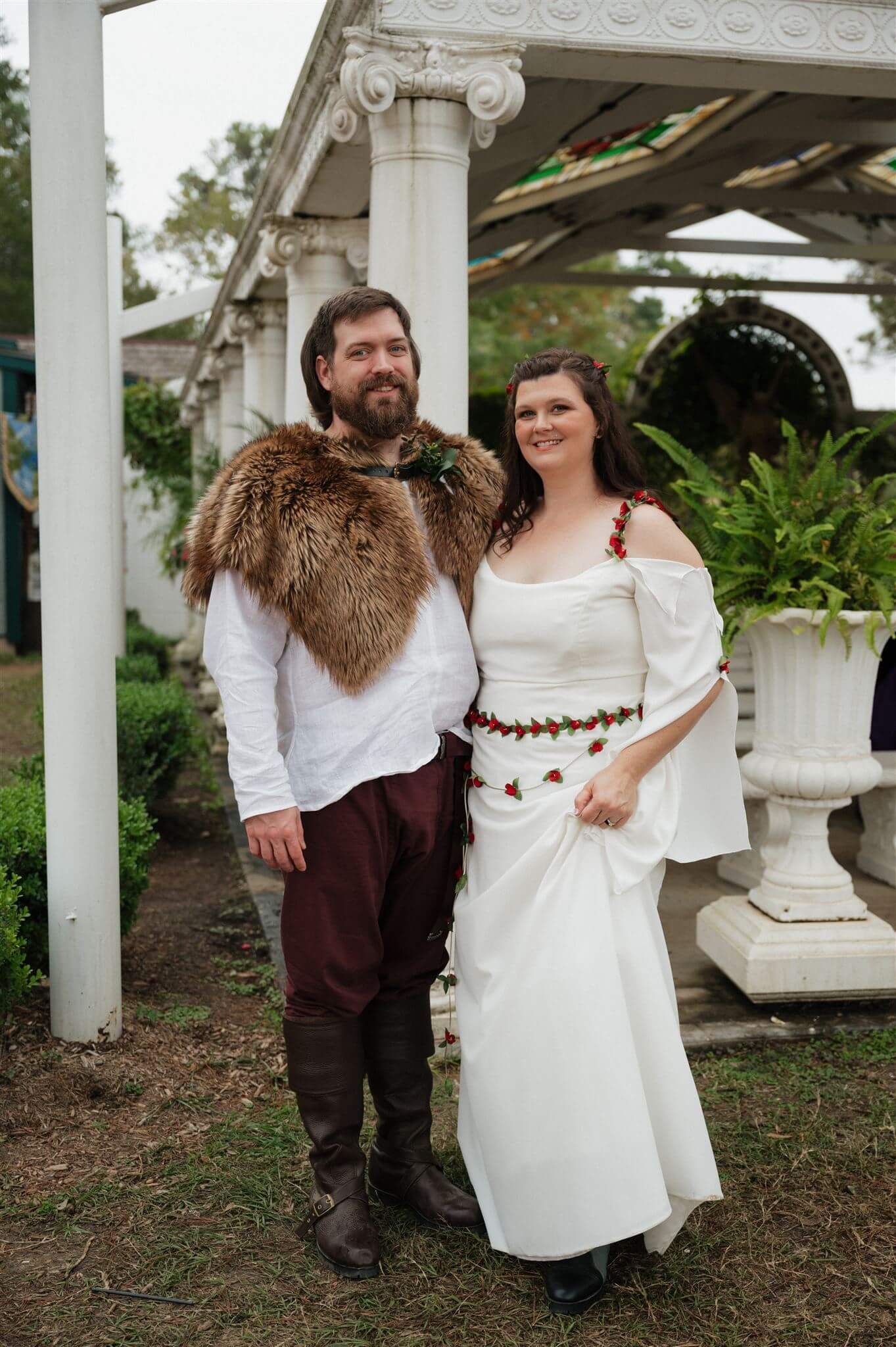 A couple in medieval-inspired wedding attire pose under a white pergola with fur cape and renaissance clothing.