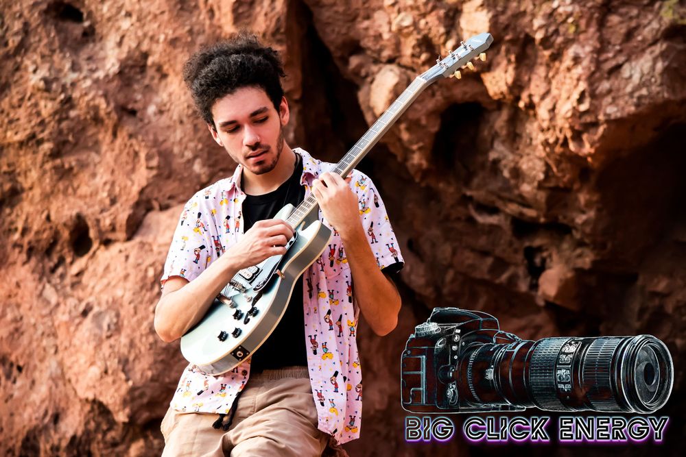 Young man with natural hair and a nose ring, posing outdoors in the desert with a cool blue tone. Playing guitar.