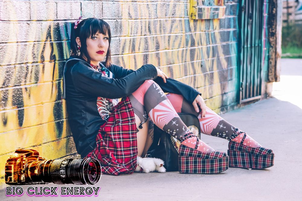 Anime-inspired model in a plaid skirt, red lipstick, and cute uwu pose, standing outdoors by graffiti in Phoenix, AZ.