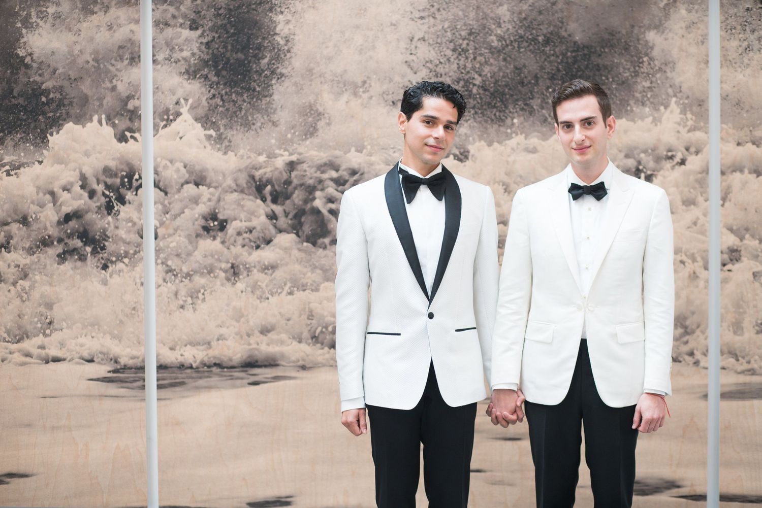 Two grooms in matching white tuxedo jackets hold hands against a desert backdrop.