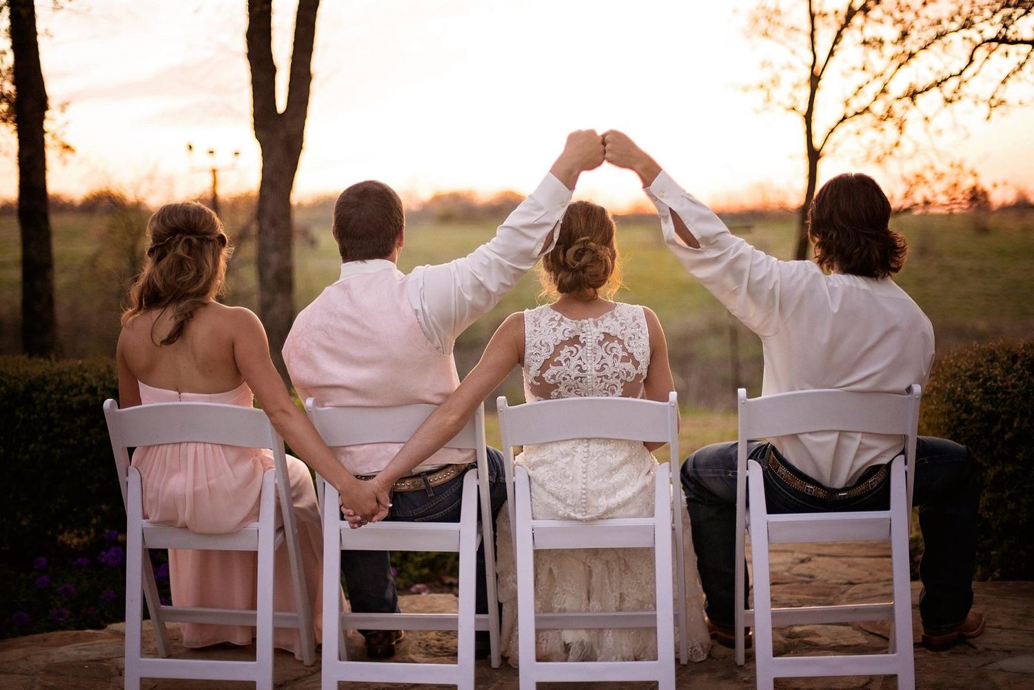 Wedding party seated in white chairs at sunset creating silhouettes against golden sky.