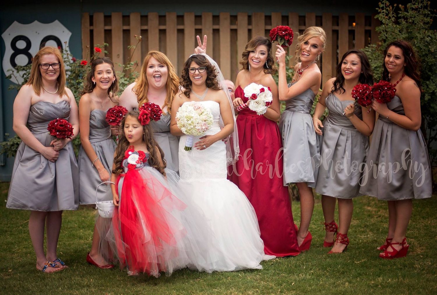 Wedding party in red and gray dresses poses together on grassy lawn at the shack in the back in Midland Texas.