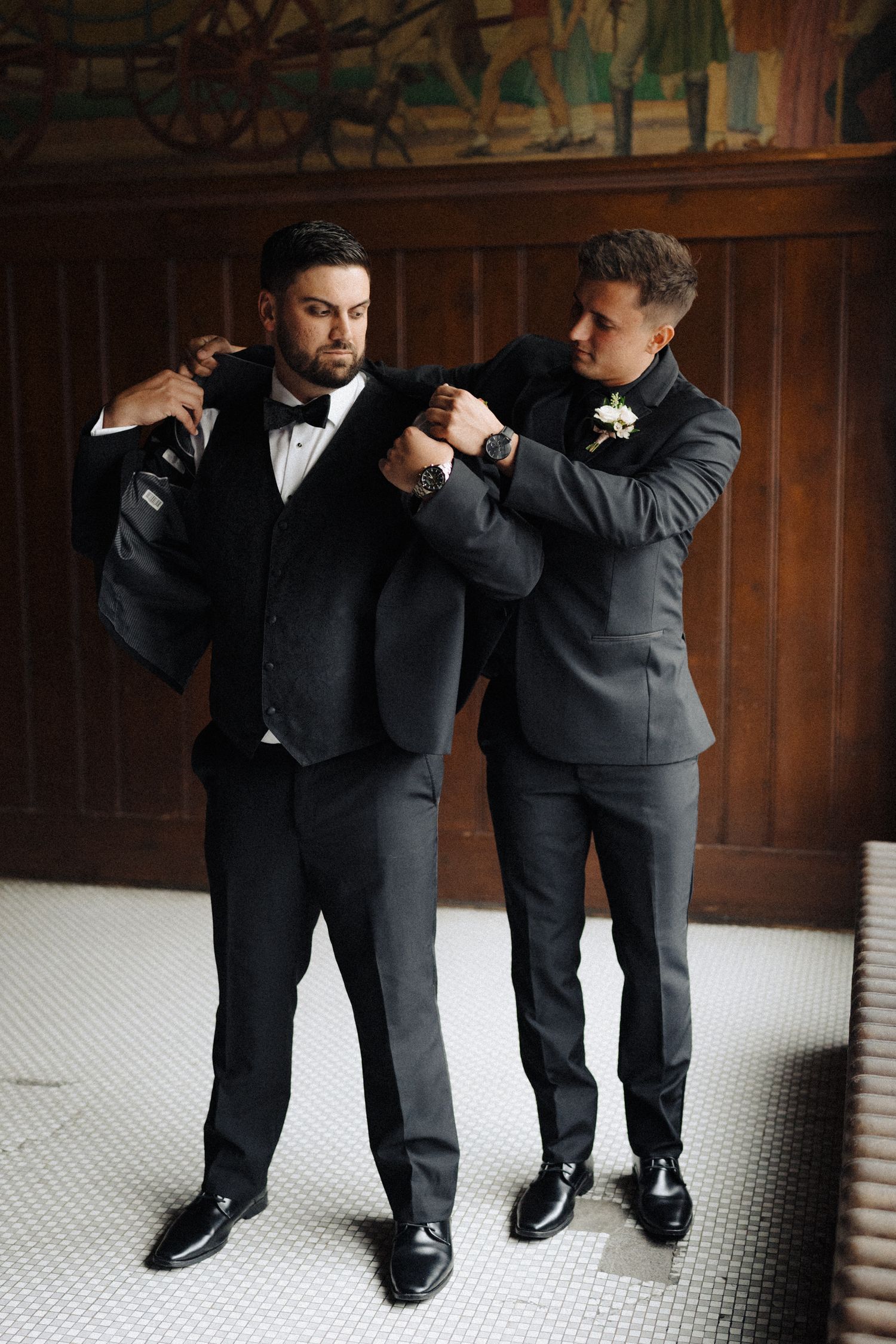 Two men in formal tuxedos playfully adjust clothing during wedding preparations.