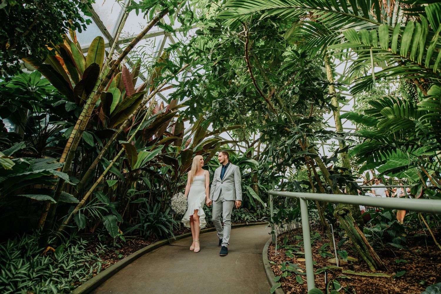 Newlyweds walk hand in hand through tropical garden pathway surrounded by palm fronds.