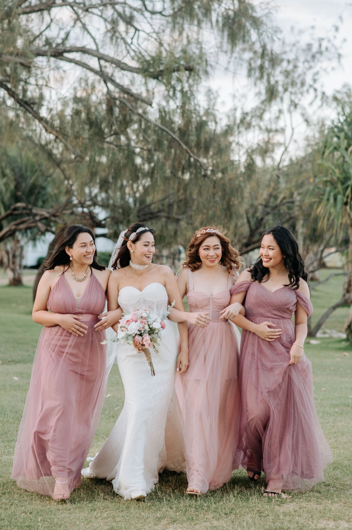Bridal party in flowing pink dresses pose together outdoors under willow trees.