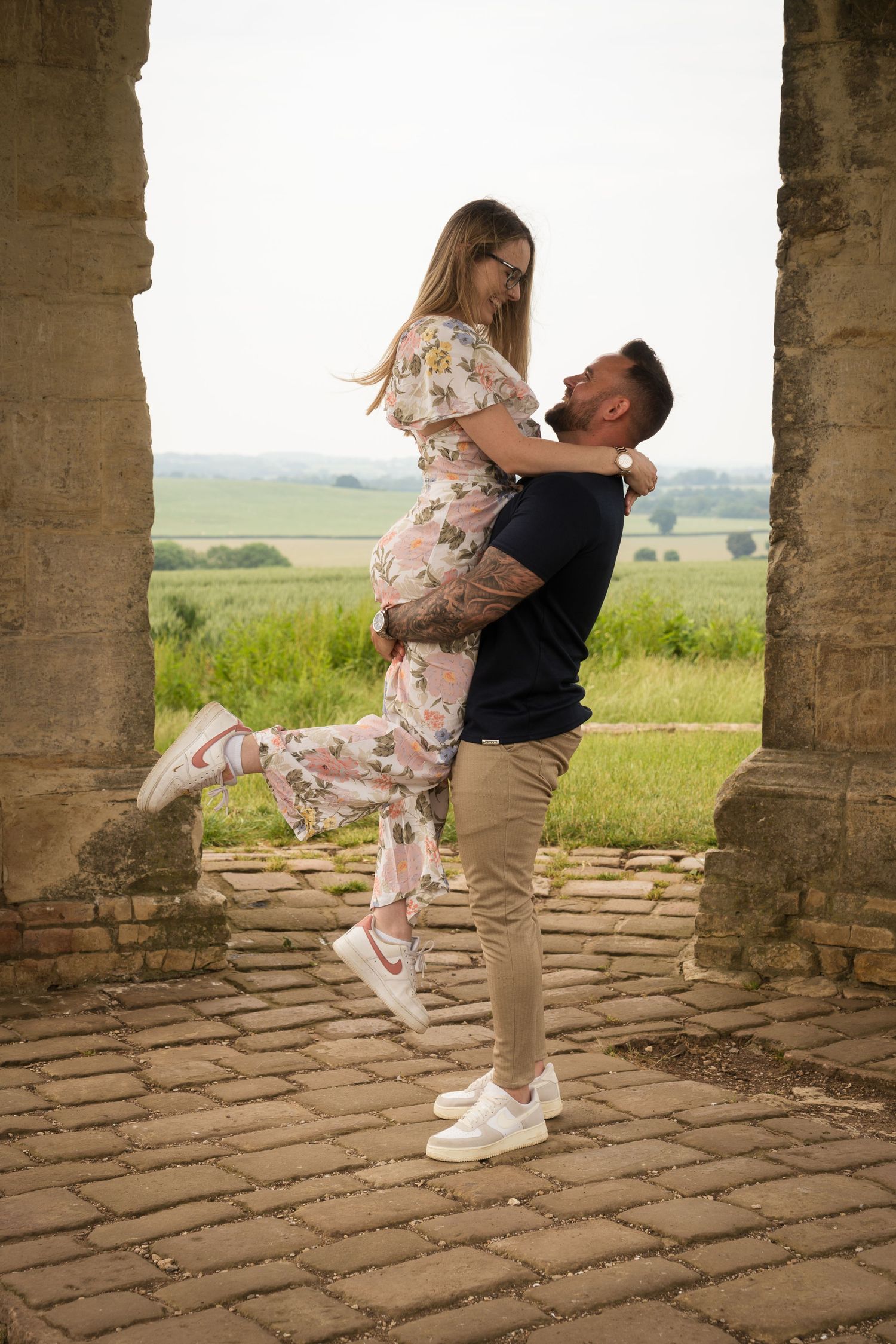A young couple, the male is lifting up the female, whilst she is popping her leg. They are looking at each other with big smiles.