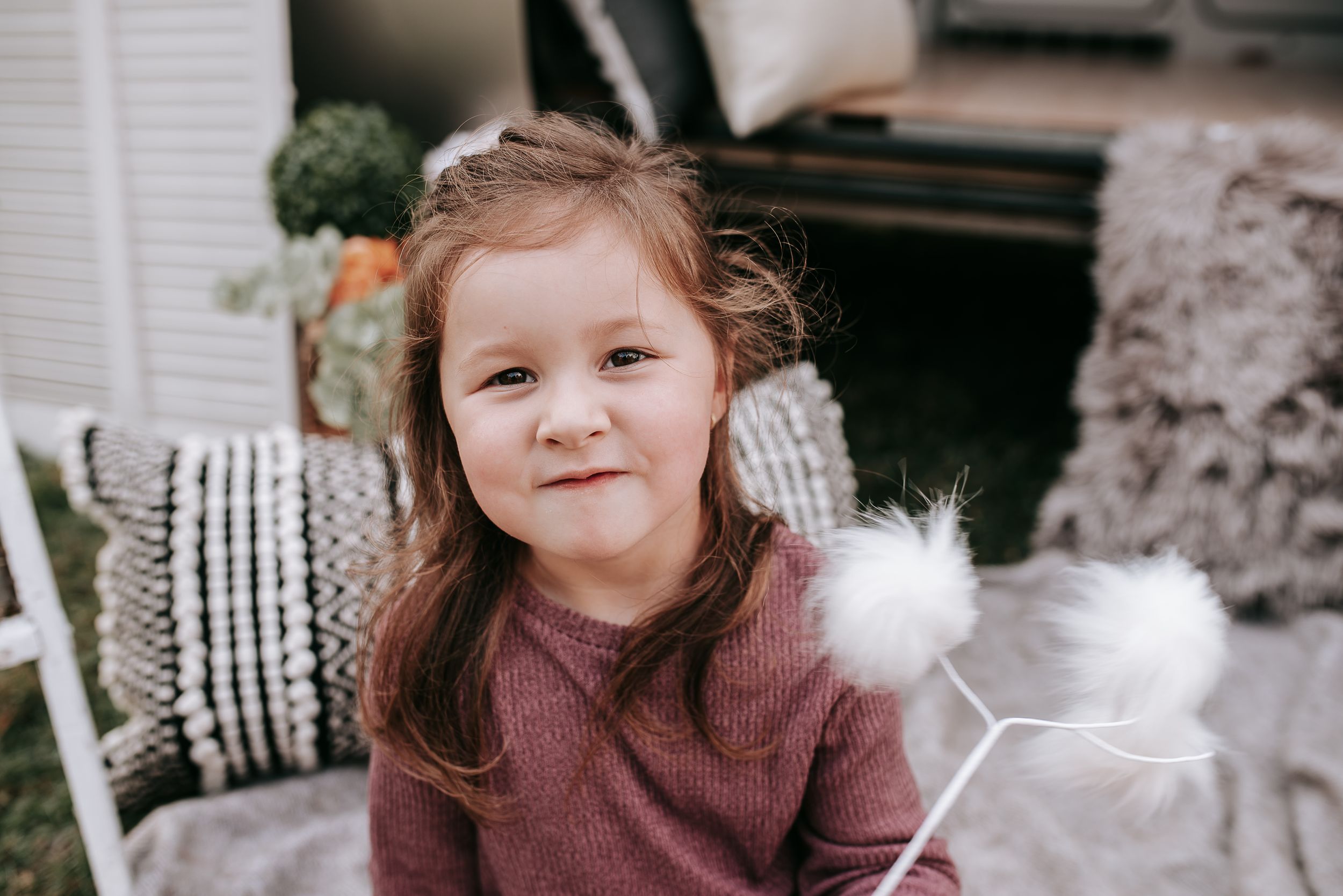 Young child wearing a maroon sweater smiling while standing next to decorative white furniture.