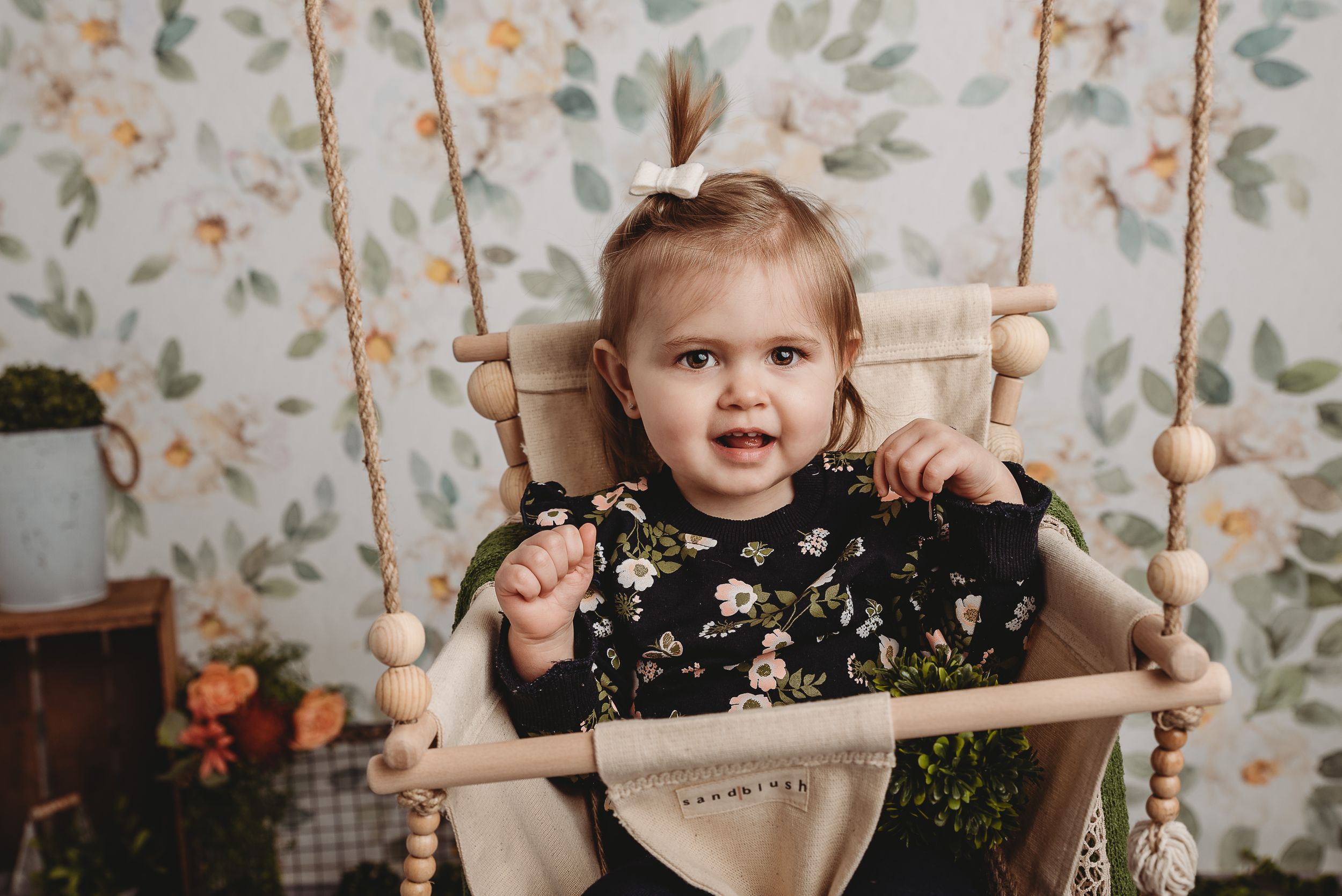 Portrait of toddler in floral dress sitting in wooden swing against vintage floral wallpaper.