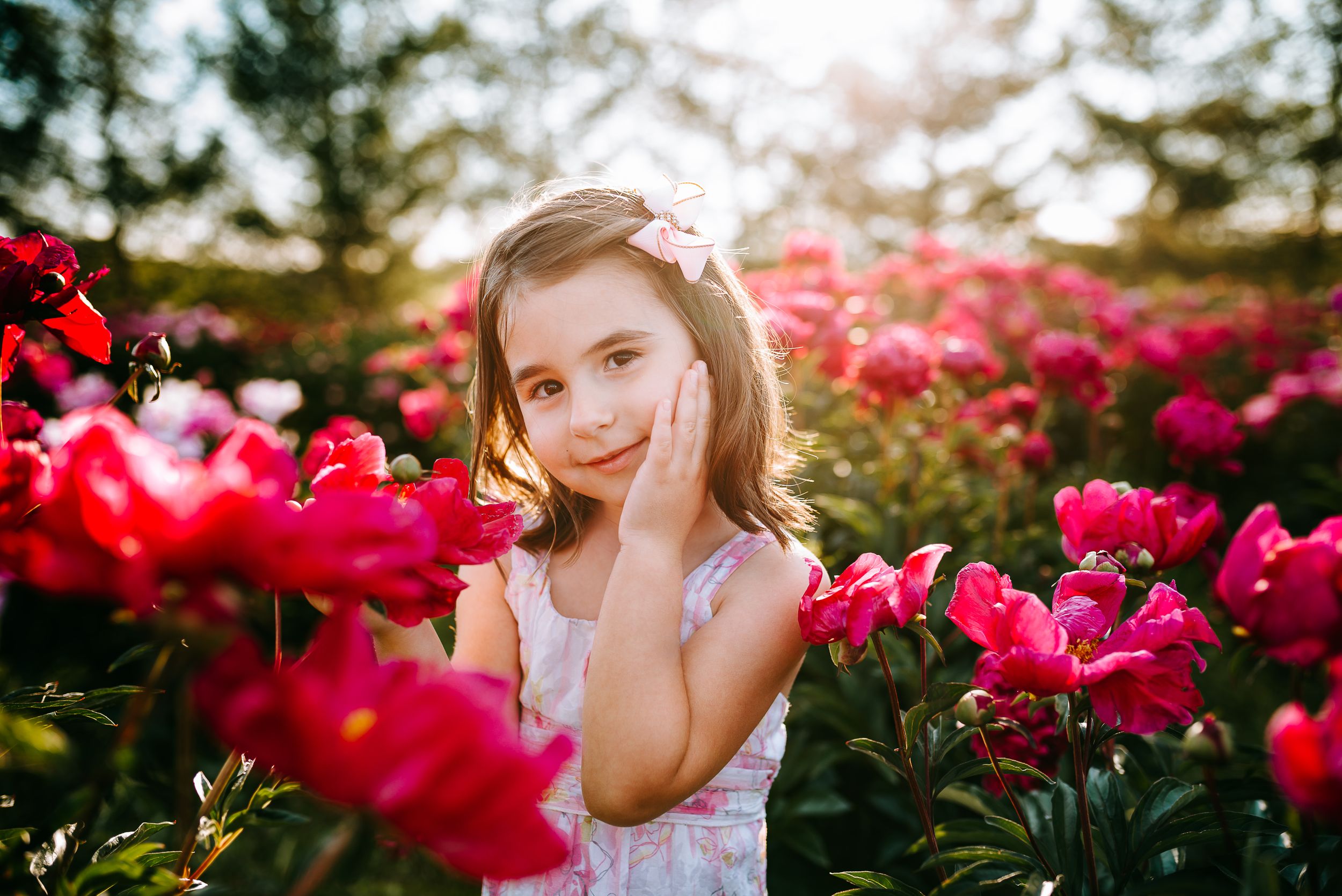 Child in white dress posing among vibrant pink peonies in garden.
