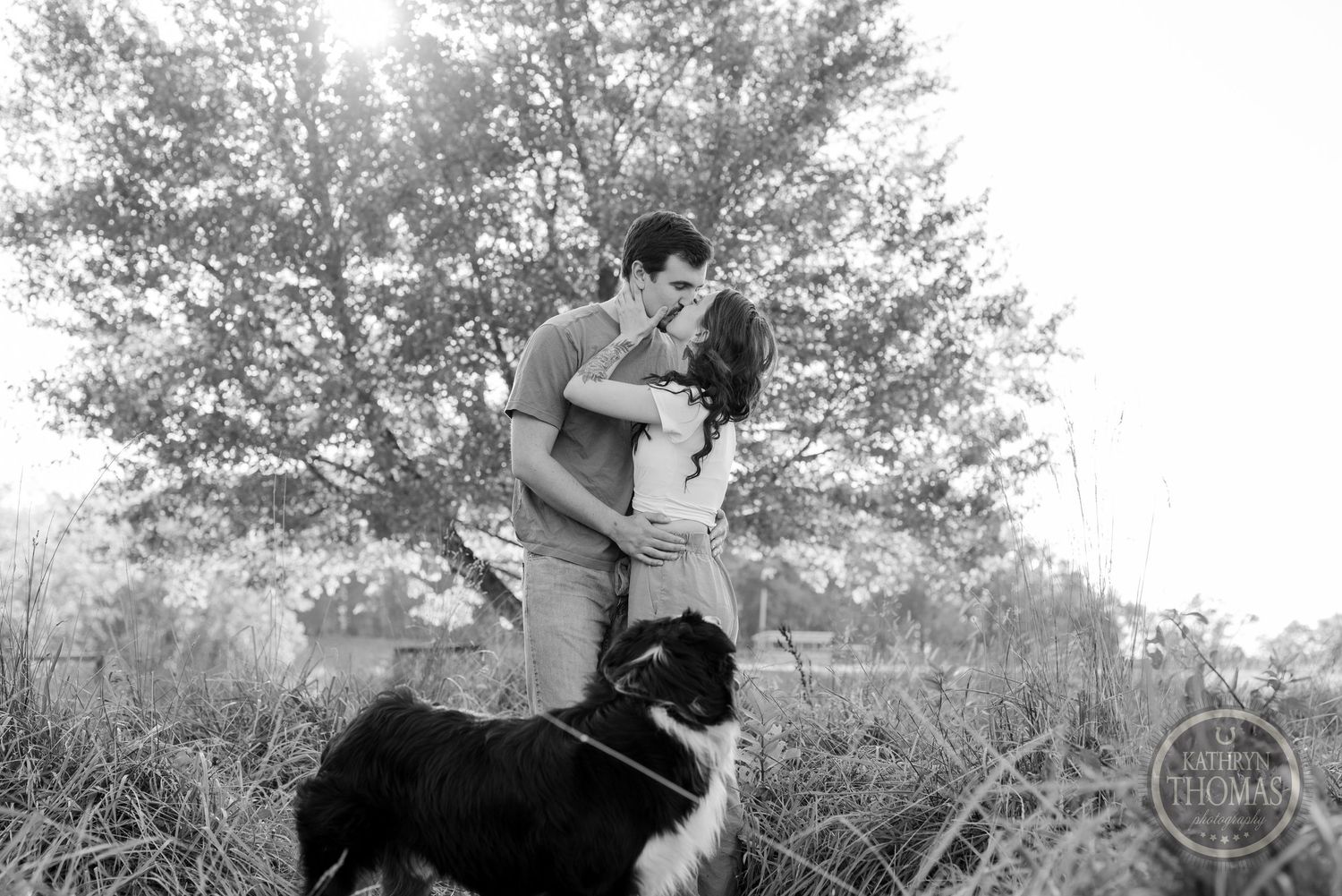 A black and white portrait of a couple embracing outdoors with their dog watching nearby.