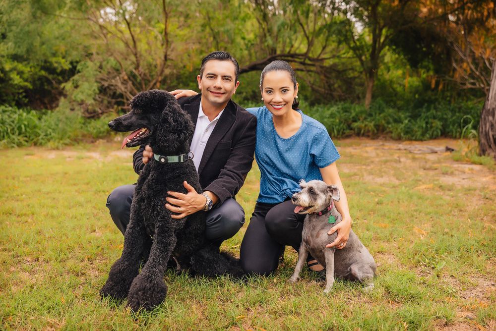 Happy couple kneels with their two dogs on grassy field surrounded by trees.