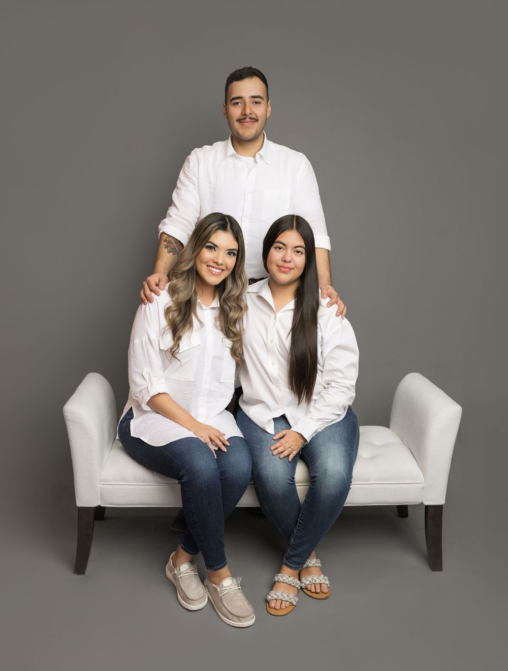 Three people in coordinated white shirts and jeans pose together on white bench in studio setting.