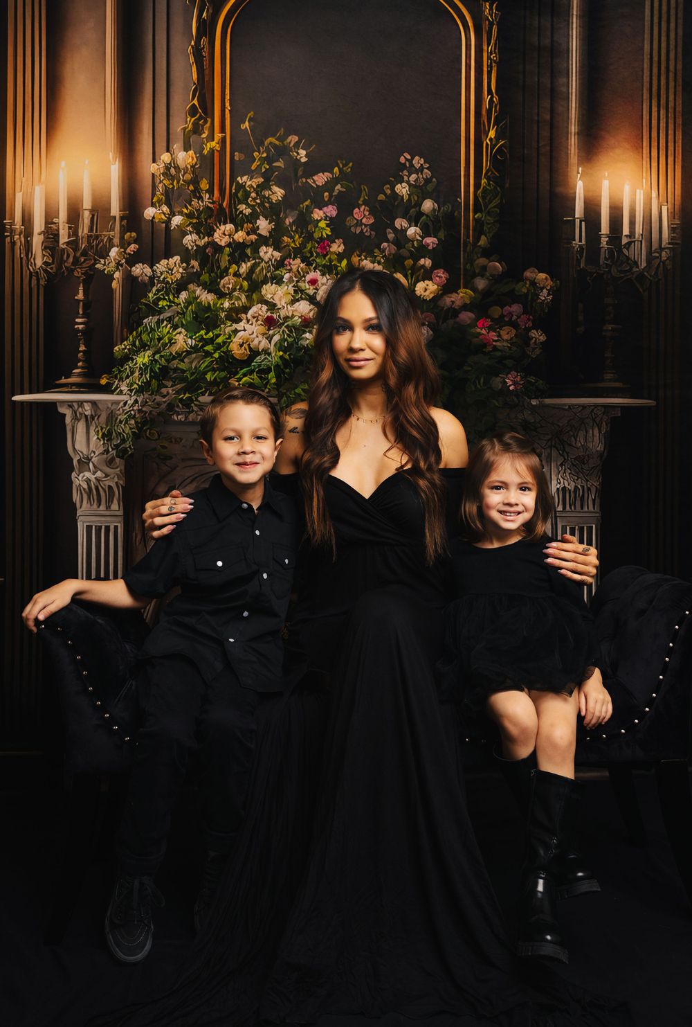 Family portrait with three people in coordinated black outfits against ornate floral and candlelit backdrop.