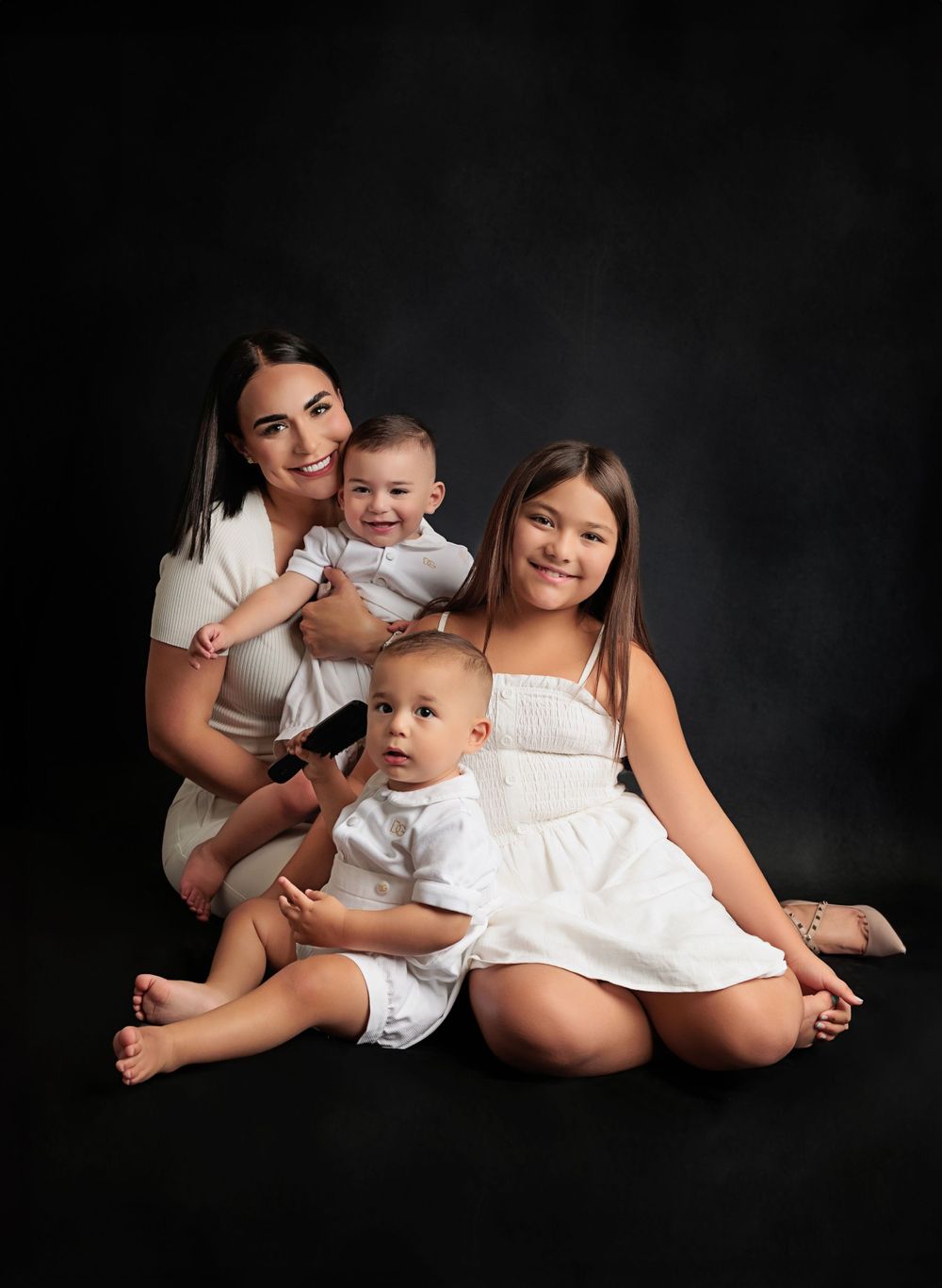 Intimate family portrait with mother and three children in white outfits against dark background.