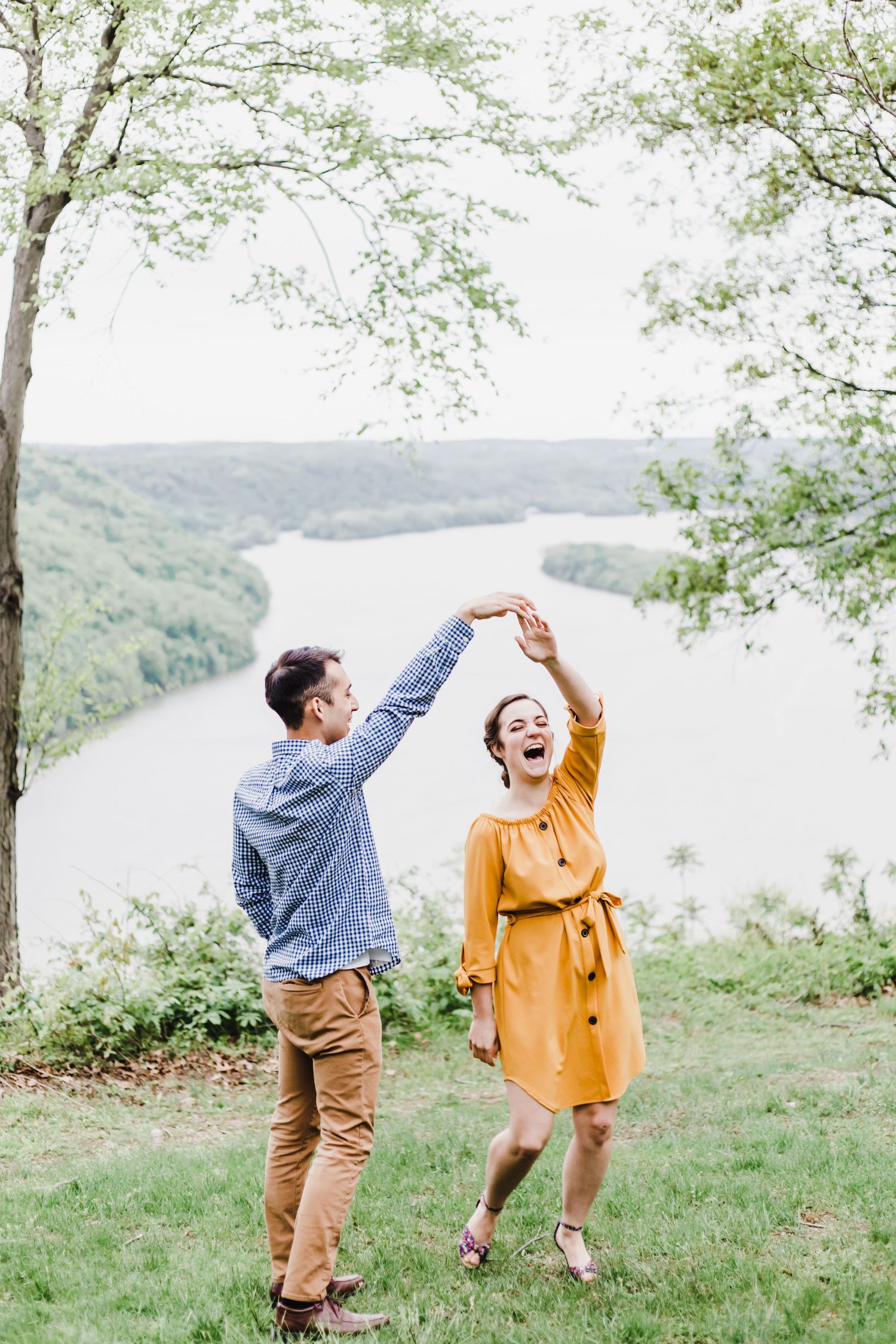 A couple dances joyfully on a hilltop overlooking a scenic river valley on a bright spring day.